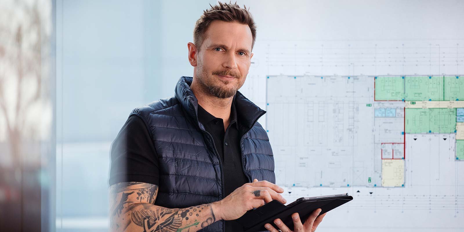 Ein Mann hält ein Tablet in der Hand und im Hintergrund ist eine Tafel an der eine Technische Zeichnung hängt