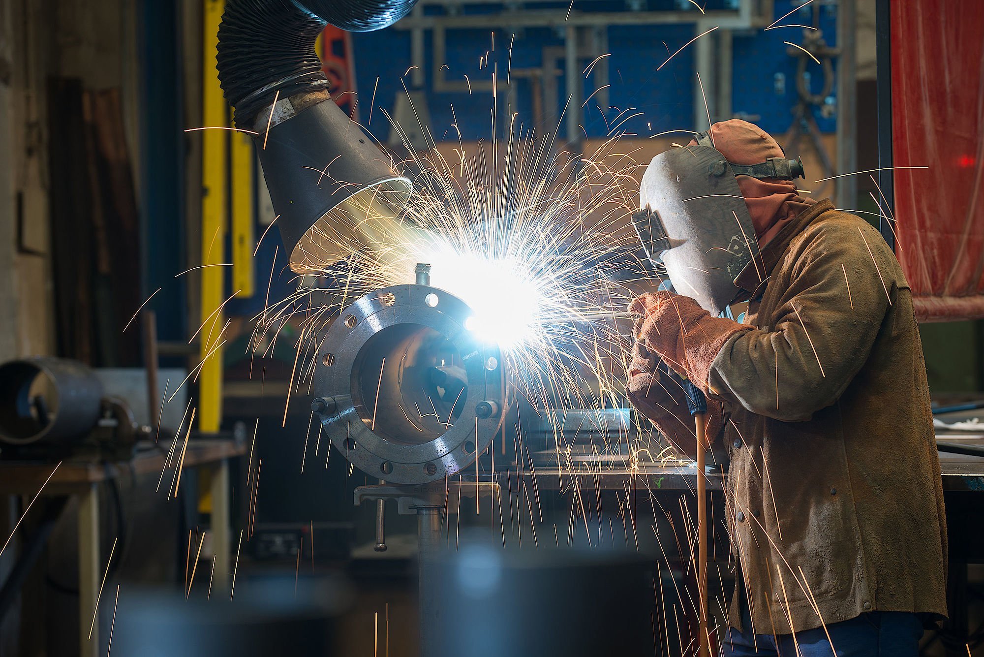 Ein Schweißer arbeitet konzentriert an einem Metallstück, während Funken in alle Richtungen sprühen und eine helle Lichtquelle entsteht.