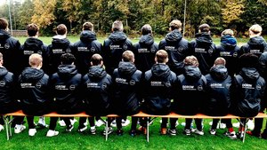 Die Spieler sind von hinten zu sehen, während sie auf einer Bank auf einem Fußballfeld sitzen, umgeben von Bäumen.