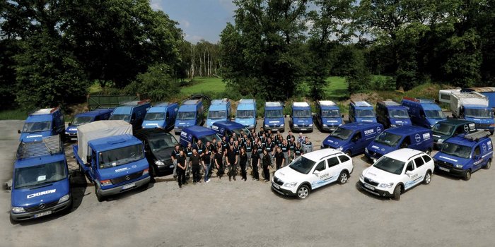 Eine Gruppe von Mitarbeitern steht vor einer Vielzahl blauer Lieferwagen auf einem Parkplatz, umgeben von Bäumen.