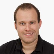 Ein junger Mann mit kurzem, braunem Haar lächelt freundlich in die Kamera, während er ein schwarzes T-Shirt trägt.