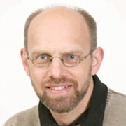 Ein Mann mit Brille und einem freundlichen Lächeln, trägt einen hellen Pullover und hat eine Glatze.