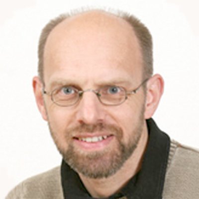 Ein Mann mit Brille und einem freundlichen Lächeln, trägt einen hellen Pullover und hat eine Glatze.