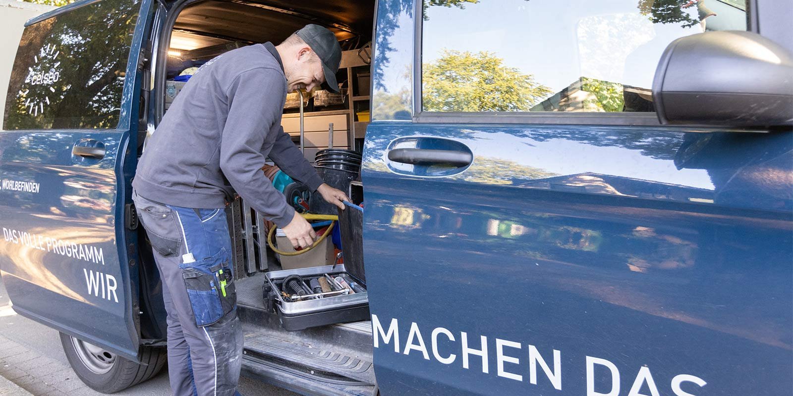 Ein Handwerker steht an einem offenen Lieferwagen und sortiert Werkzeuge aus einer Kiste, während er lächelt.