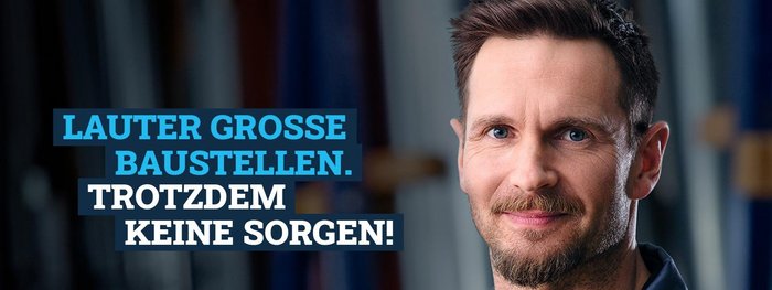 Ein lächelnder Mann mit Bart steht vor einem unscharfen Hintergrund, der Baustellen und Materialien zeigt.