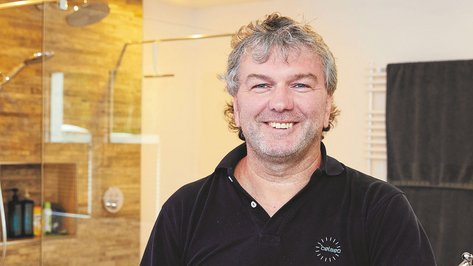 Ein lächelnder Mann mit grauen Haaren steht in einem modernen Badezimmer mit Holzverkleidung und Handtüchern im Hintergrund.