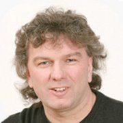 Ein Mann mit lockigem, grauem Haar lächelt freundlich in die Kamera, während er ein schwarzes Oberteil trägt.