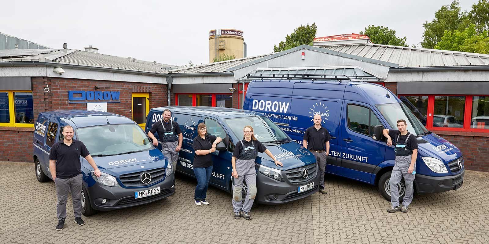 Die Gruppe von Technikern in Arbeitskleidung posiert vor zwei blauen Lieferwagen, die für Sanitär- und Heizungsdienstleistungen genutzt werden.