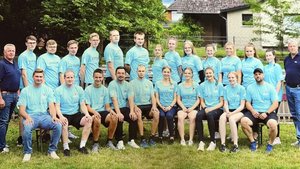 Ein Team von jungen Erwachsenen in hellblauen Shirts steht vor einem Haus, umgeben von Bäumen und Rasen.